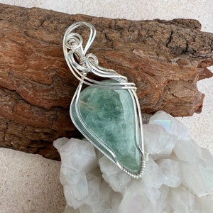 May include: A silver wire wrapped pendant featuring a light green gemstone. The pendant is displayed on a piece of driftwood and a bed of white crystals.