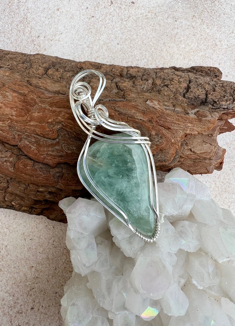 May include: A silver wire wrapped pendant featuring a light green gemstone. The pendant is displayed on a piece of driftwood and a bed of white crystals.