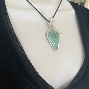May include: A silver wire wrapped pendant necklace featuring a light green gemstone. The necklace is on a black cord.