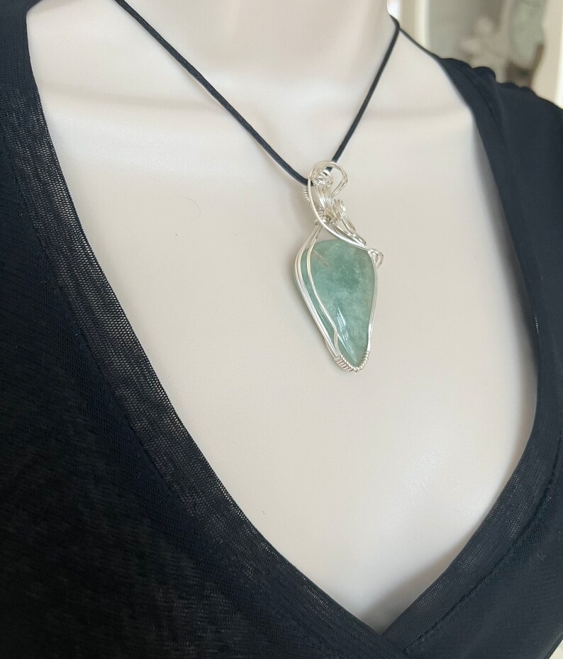 May include: A silver wire wrapped pendant necklace featuring a light green gemstone. The necklace is on a black cord.