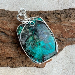 Puede incluir: Un colgante de piedra preciosa verde y negra con envoltura de alambre plateado. El colgante est&aacute; sobre un trozo de madera a la deriva.