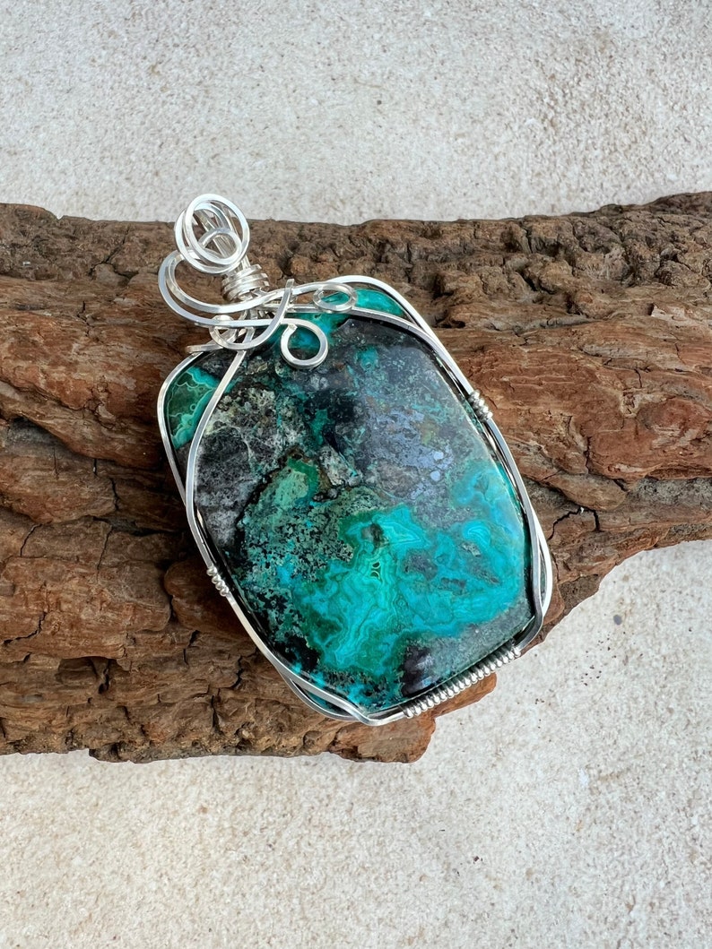 Puede incluir: Un colgante de piedra preciosa verde y negra con envoltura de alambre plateado. El colgante est&aacute; sobre un trozo de madera a la deriva.