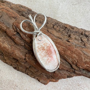 May include: A silver wire wrapped pendant featuring a pink and white agate stone. The wire is shaped into a heart at the top of the pendant.