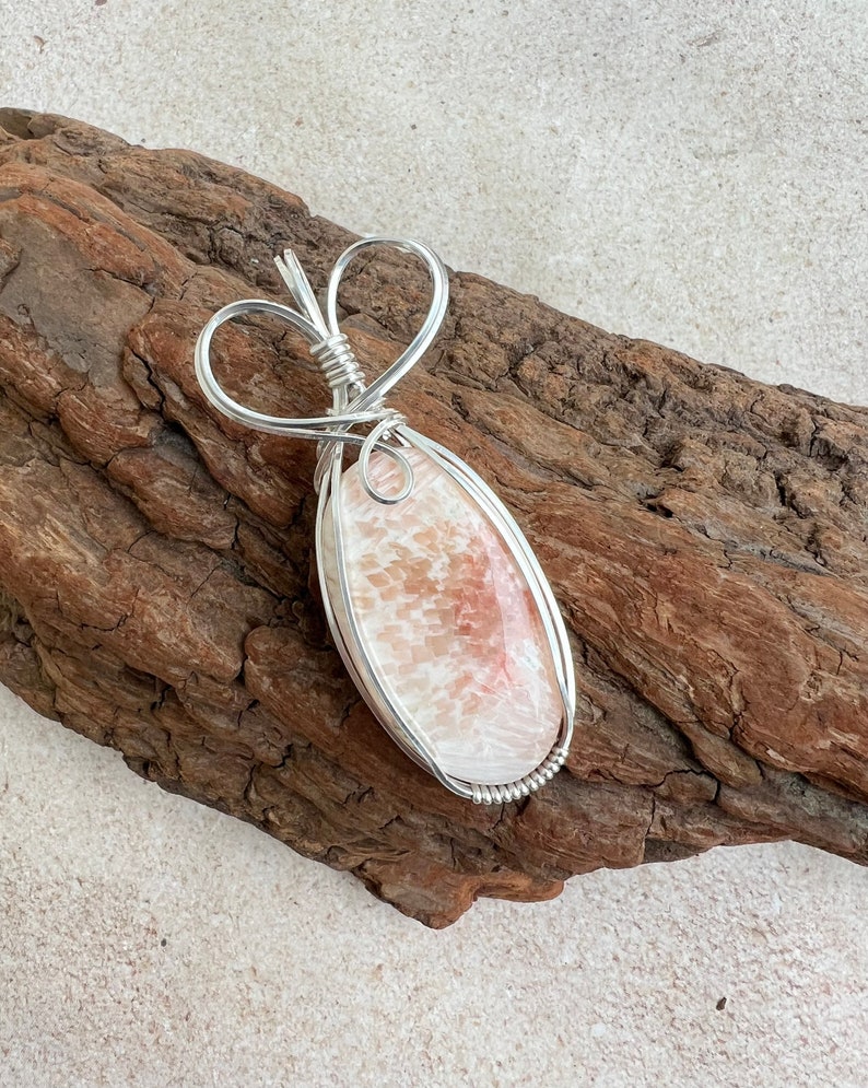 May include: A silver wire wrapped pendant featuring a pink and white agate stone. The wire is shaped into a heart at the top of the pendant.