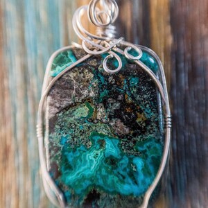 Puede incluir: Un colgante de alambre plateado con una piedra preciosa verde, negra y dorada. La piedra preciosa tiene un patr&oacute;n veteado &uacute;nico.