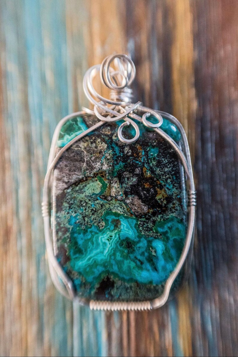 Puede incluir: Un colgante de alambre plateado con una piedra preciosa verde, negra y dorada. La piedra preciosa tiene un patr&oacute;n veteado &uacute;nico.