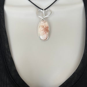 May include: A silver wire wrapped pendant featuring a light brown and white oval stone with a delicate heart-shaped bail. The pendant is hanging from a black cord necklace.