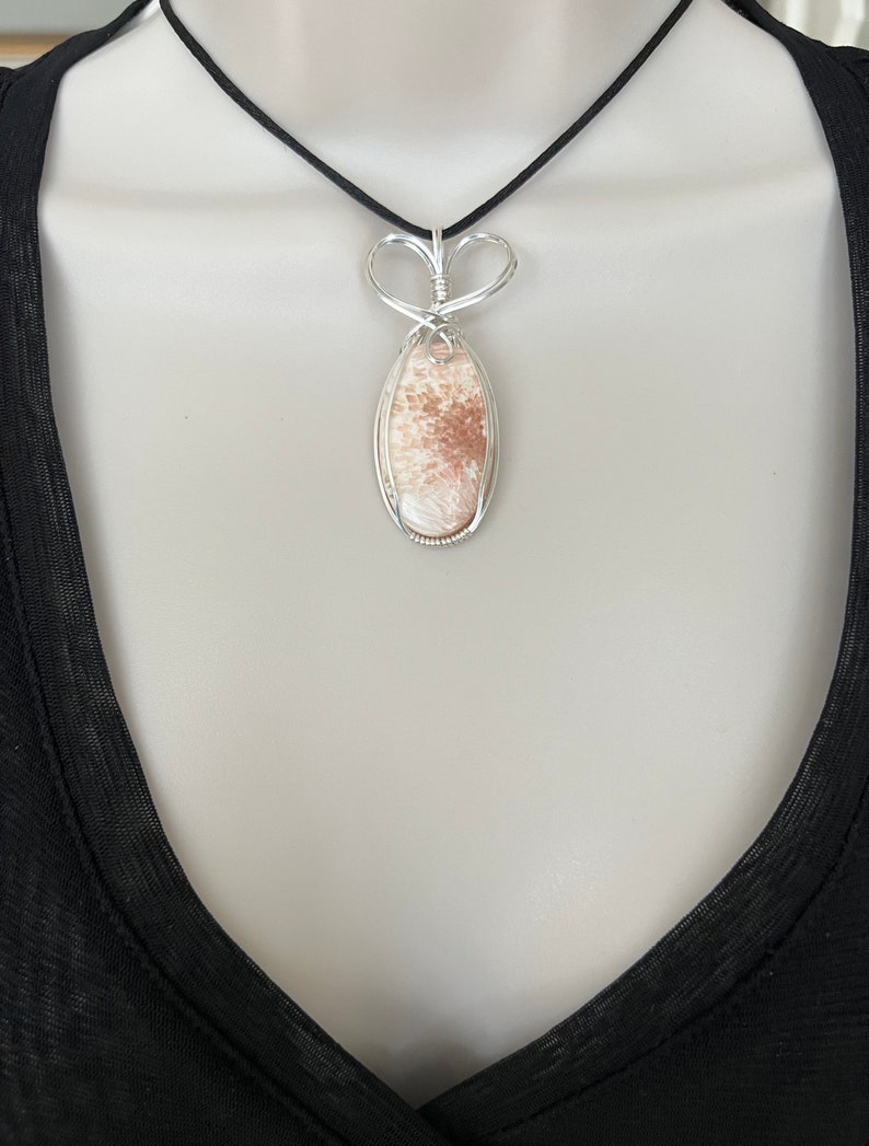 May include: A silver wire wrapped pendant featuring a light brown and white oval stone with a delicate heart-shaped bail. The pendant is hanging from a black cord necklace.