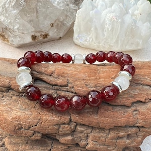 May include: A red gemstone bracelet with silver beads and white crystal accents. The bracelet is on a piece of wood.