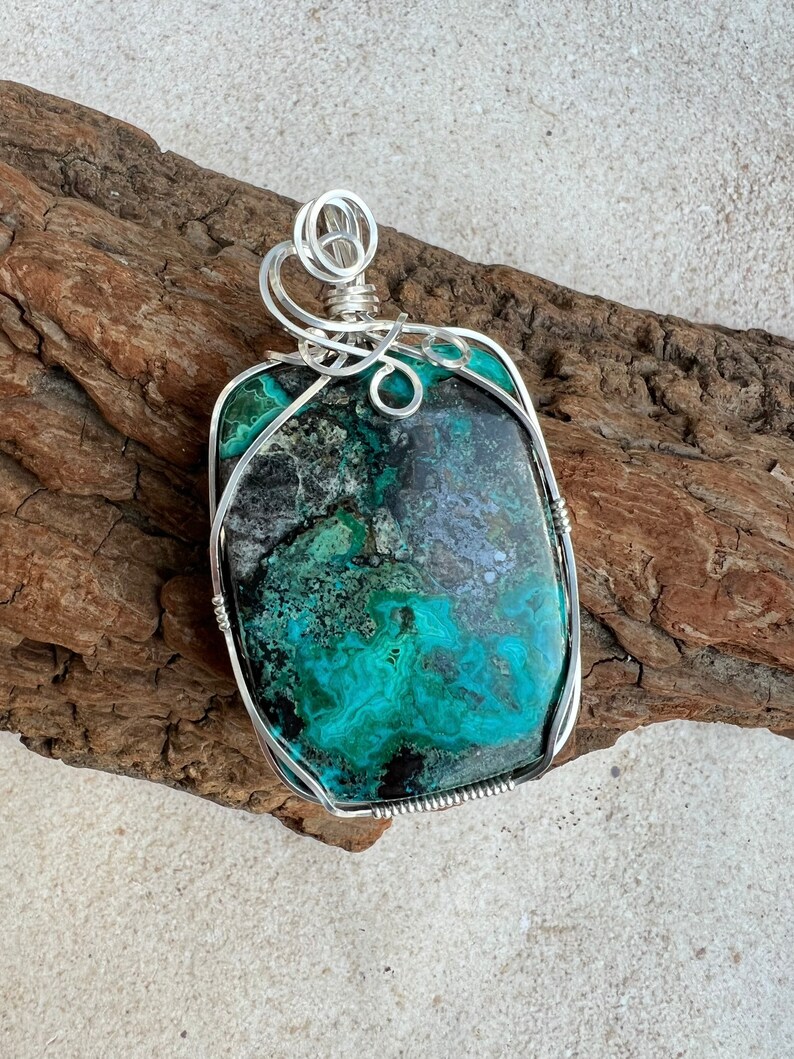 Puede incluir: Un colgante de alambre plateado envuelto alrededor de una piedra grande de crisocola verde y negra. La piedra tiene un patr&oacute;n &uacute;nico y arremolinado.