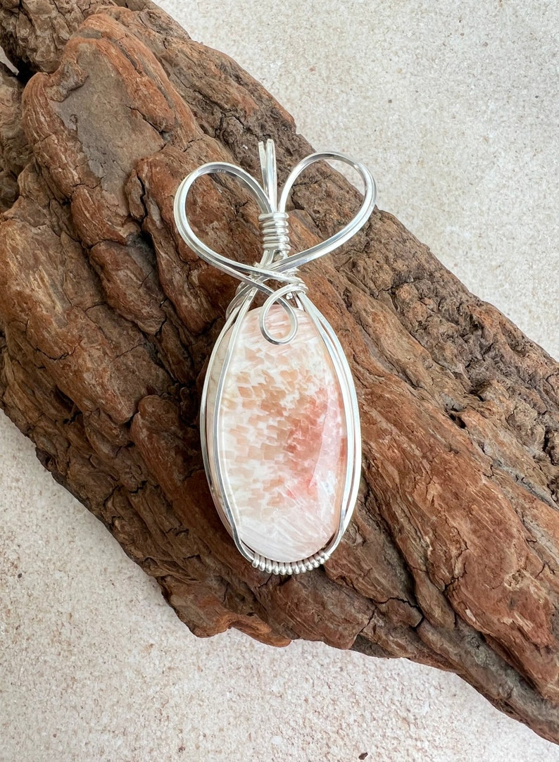 May include: A silver wire wrapped pendant featuring a pink and white oval shaped stone with a heart shaped bail.