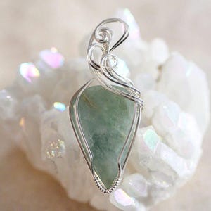 May include: A silver wire wrapped pendant featuring a light green gemstone. The pendant is displayed on a bed of white crystals.