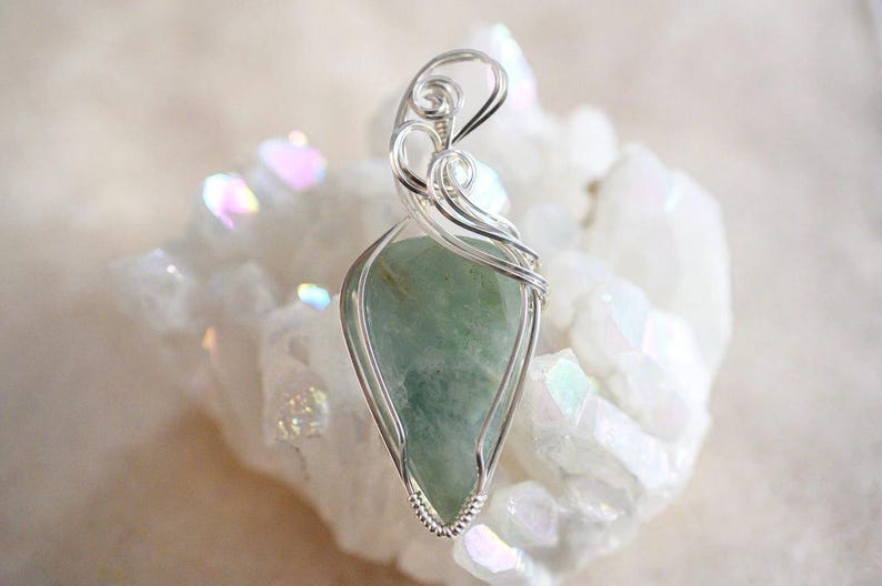 May include: A silver wire wrapped pendant featuring a light green gemstone. The pendant is displayed on a bed of white crystals.