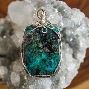 Puede incluir: Un colgante de piedra preciosa verde y negra con envoltura de alambre plateado. El colgante est&aacute; sobre un lecho de cristales blancos.