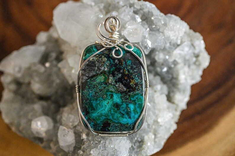 Puede incluir: Un colgante de piedra preciosa verde y negra con envoltura de alambre plateado. El colgante est&aacute; sobre un lecho de cristales blancos.
