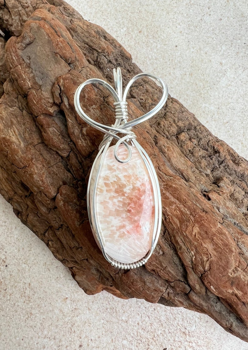 May include: A silver wire wrapped pendant featuring a pink and white agate stone. The pendant is shaped like a heart and has a delicate design.