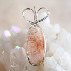 May include: A delicate silver wire wrapped pendant featuring a light pink and white coral stone. The pendant is shaped like a heart and is set against a white background.