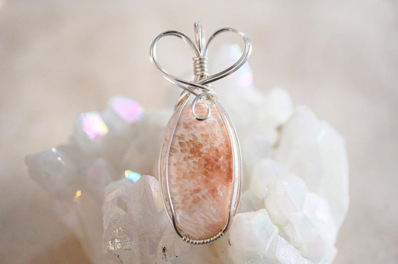 May include: A delicate silver wire wrapped pendant featuring a light pink and white coral stone. The pendant is shaped like a heart and is set against a white background.