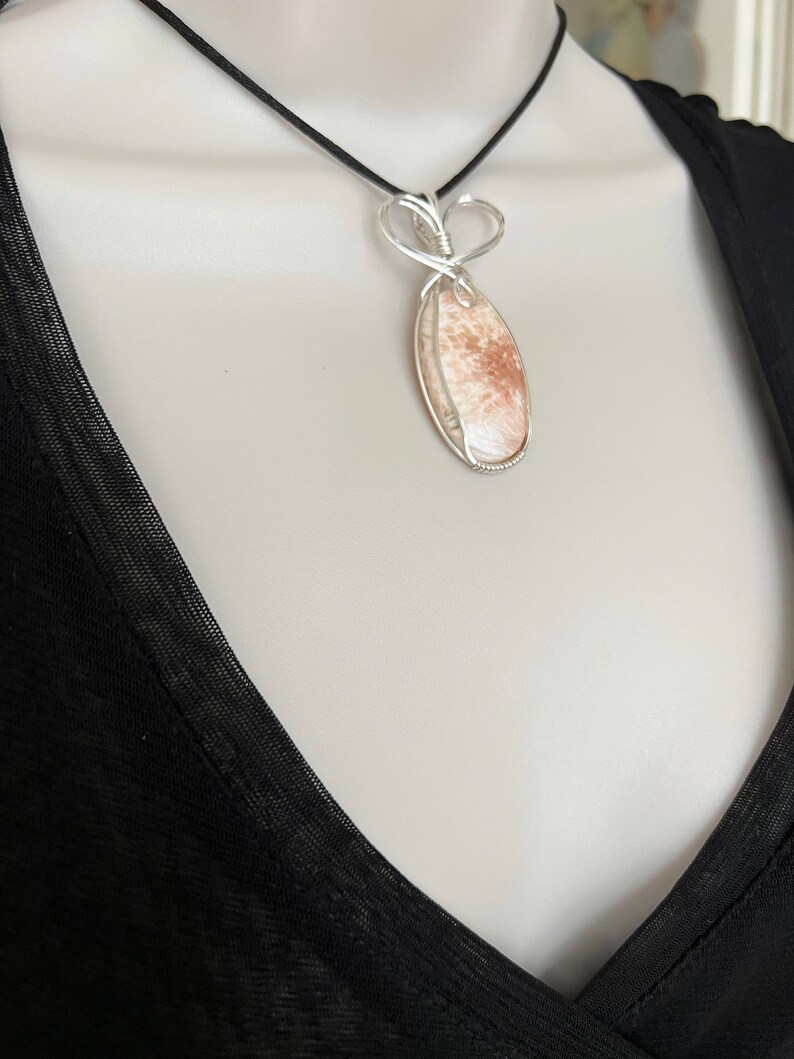 May include: A silver wire wrapped pendant necklace featuring a light brown and white stone with a heart-shaped bail.