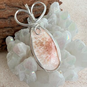 May include: A silver wire wrapped pendant featuring a pink and white agate stone. The pendant is shaped like a teardrop and is set on a bed of white crystals.