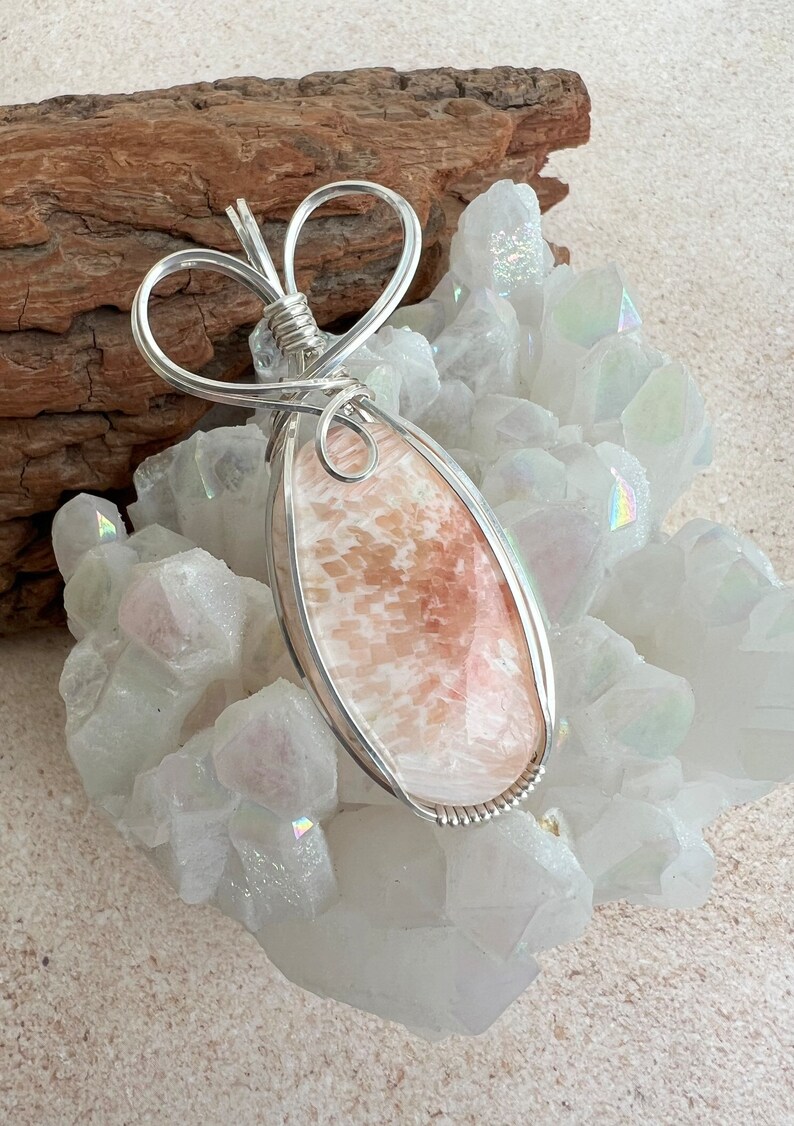 May include: A silver wire wrapped pendant featuring a pink and white agate stone. The pendant is shaped like a teardrop and is set on a bed of white crystals.