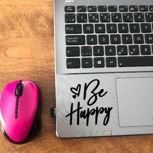 Peut inclure: Un ordinateur portable argenté avec un clavier noir et une souris noire. L'ordinateur portable a un autocollant sur le pavé tactile qui dit "Be Happy" avec un symbole de cœur.