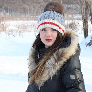 May include: A woman wearing a gray knit hat with a red and white stripe and a fur-trimmed black winter jacket. She is standing in a snowy landscape.