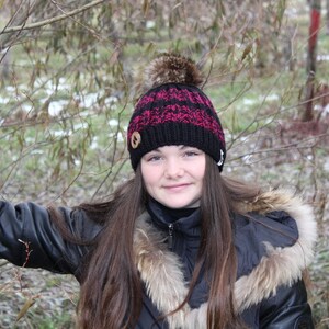 Peut inclure: Une jeune personne aux longs cheveux bruns portant un bonnet en tricot rayé noir et rouge avec un pompon brun sur le dessus. Le bonnet a un bouton en bois sur le devant. La personne porte une veste en cuir noir avec un col en fourrure.