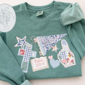 May include: Teal sweatshirt with appliqued cross, star, heart, and hand-shaped designs. The sweatshirt features a floral banner and a book with the text "Psalm 23:1-4". The design includes a floral logo in the upper left corner.