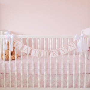 May include: A white crib with a pink and white gingham crib sheet and a pink and white floral crib skirt. A pink banner with the name "KATHRYN" hangs above the crib.