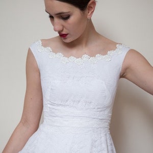 May include: A white lace dress with a scoop neckline and a fitted bodice. The dress has a full skirt and is decorated with delicate lace trim. The model is wearing a silver headband with floral accents.