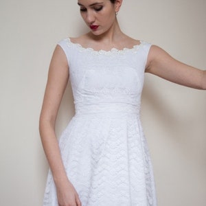 May include: A white lace dress with a scoop neckline and a fitted bodice. The dress has a full skirt and is adorned with a delicate floral pattern. The model is wearing a silver headband with floral accents.
