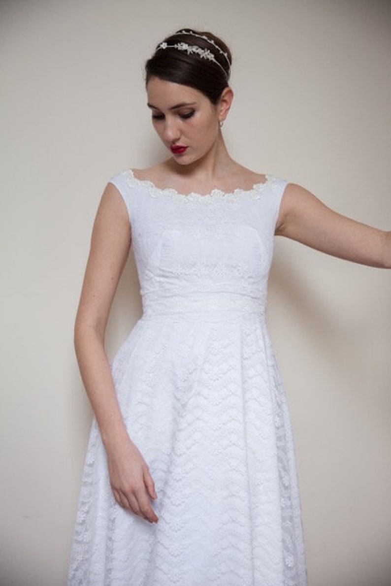 May include: A white lace dress with a scoop neckline and a fitted bodice. The dress has a full skirt and is adorned with a delicate floral pattern. The model is wearing a silver headband with floral accents.
