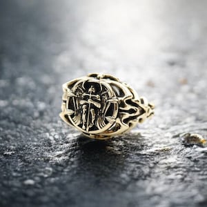 May include: A gold ring with an intricate design featuring a winged figure holding a sword. The ring is on a dark gray surface.