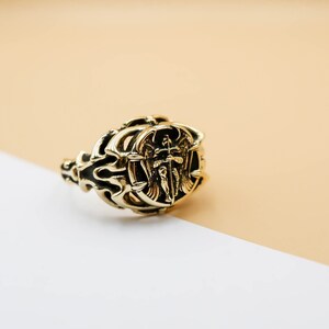 May include: A gold ring with an intricate design featuring an angel with wings holding a sword. The ring is on a white and beige surface.