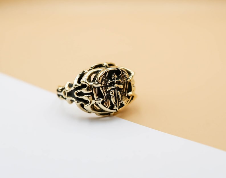 May include: A gold ring with an intricate design featuring an angel with wings holding a sword. The ring is on a white and beige surface.