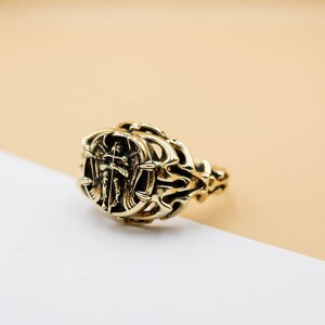 May include: A gold ring with an intricate design featuring an angel with wings holding a cross. The ring is on a light brown and white surface.