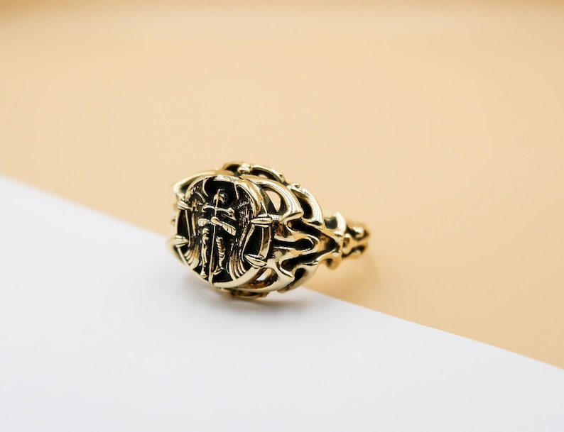 May include: A gold ring with an intricate design featuring an angel with wings holding a cross. The ring is on a light brown and white surface.