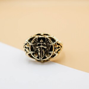 May include: A gold ring with an ornate design featuring an angel with wings holding a sword. The ring is on a beige and white surface.