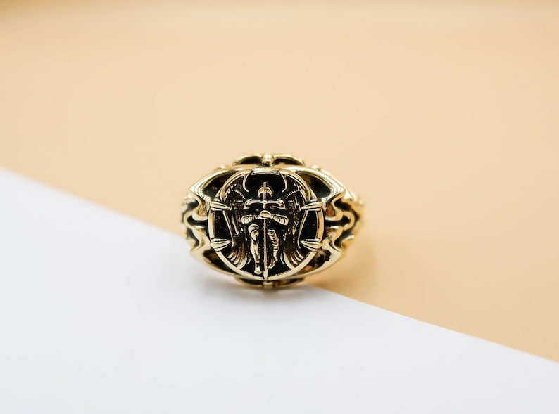 May include: A gold ring with an ornate design featuring an angel with wings holding a sword. The ring is on a beige and white surface.