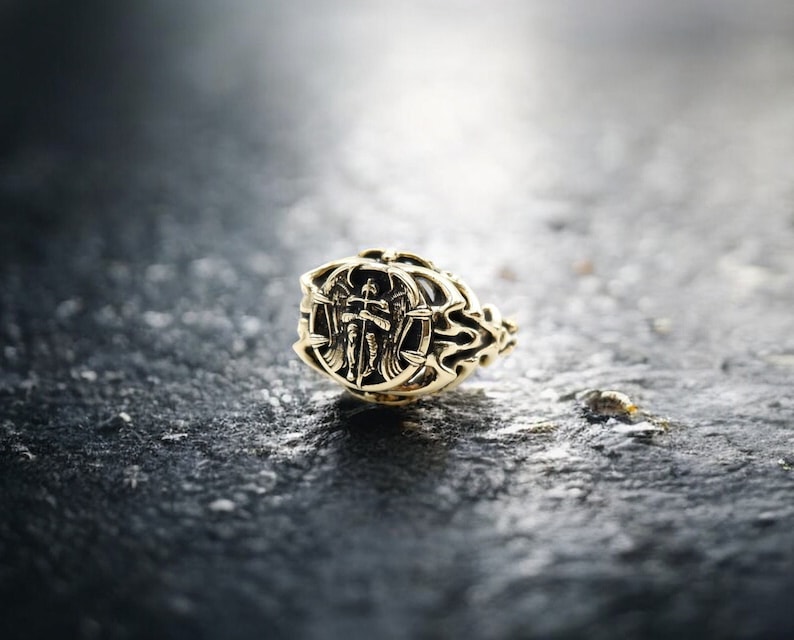 May include: A gold ring with an intricate design featuring a winged figure holding a sword. The ring is on a dark gray surface.