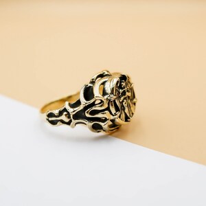 May include: A gold ring with an intricate design featuring a stylized figure. The ring is on a light brown and white background.