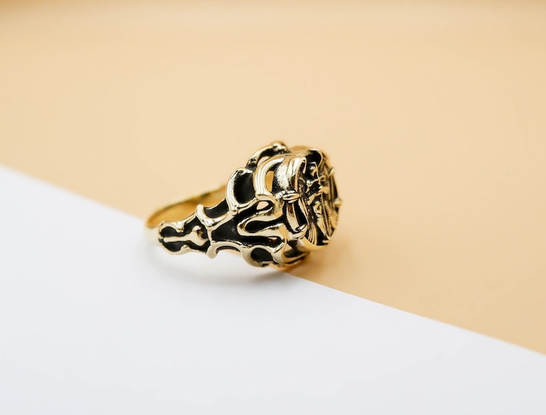 May include: A gold ring with an intricate design featuring a stylized figure. The ring is on a light brown and white background.