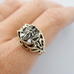 May include: A gold-toned ring with an intricate design featuring a winged creature and a shield. The ring is worn on a person's finger.