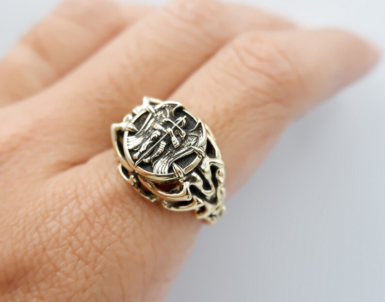 May include: A gold-toned ring with an intricate design featuring a winged creature and a shield. The ring is worn on a person's finger.
