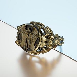May include: A gold ring with an intricate design featuring an angel with wings and a sword. The angel is set against a black background.