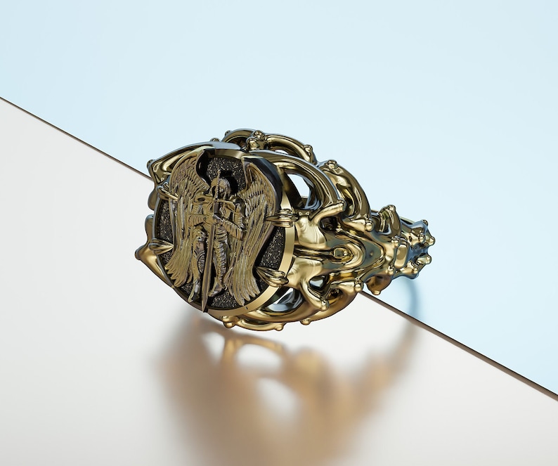 May include: A gold ring with an intricate design featuring an angel with wings and a sword. The angel is set against a black background.