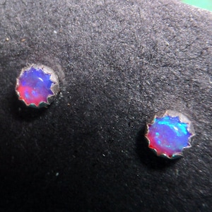 Pode incluir: Um par de brincos de prata com pequenas pedras de opala redondas e iridescentes. As pedras têm uma cor azul, roxa e rosa.