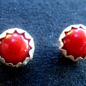 May include: A pair of stud earrings featuring a vibrant red gemstone set in a silver-toned, scalloped bezel. The earrings are round and have a polished, glossy finish, set against a dark background.