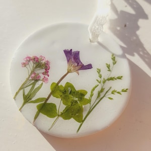 May include: A white round ornament with pressed flowers and leaves. The ornament features a purple flower, pink flowers, and green leaves.